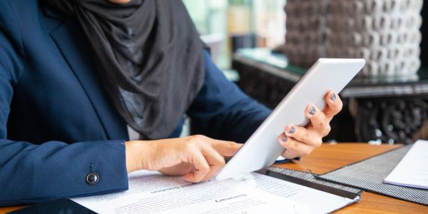 Businesswoman reading contract and consulting internet. Young Muslim business woman sitting in cafe, reading papers and using tablet. Digital communication concept